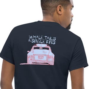 May include: Navy blue t-shirt with a pink and white graphic of a car and the text "WHALE TAILS & SINGLE FINS". The shirt is made of a soft material.
