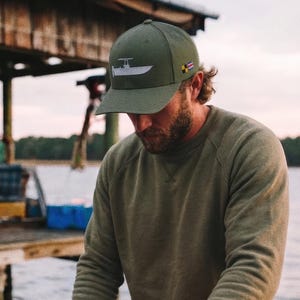 May include: An olive green cap with a boat graphic and a matching olive green sweatshirt. An orange basket filled with shrimp sits on a wooden dock. The person is sorting shrimp.