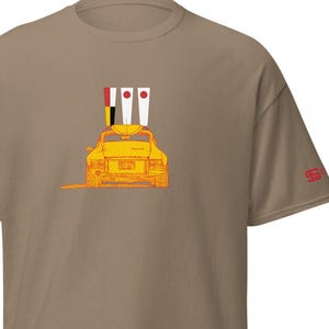 May include: A tan t-shirt featuring a yellow sports car graphic with three flags on top. The car is outlined in orange, and the flags have red and white details. The sleeve has a small red logo.