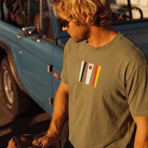 May include: Olive green t-shirt with a graphic design of vertical stripes in black, white, red, and yellow. The t-shirt is being worn near a blue vintage car and a brown duffel bag.