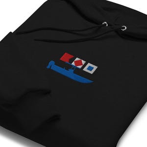 May include: Black hoodie featuring an embroidered design of a blue boat with a white motor, red and white flags, and a white square. The sweatshirt has a drawstring hood and a front pocket. The design is nautical themed.