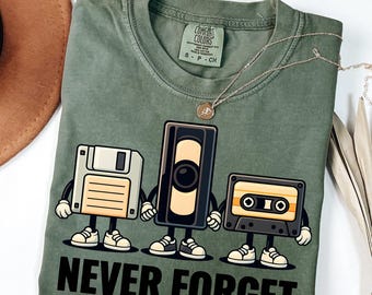 May include: A moss green t-shirt featuring a graphic of a floppy disk, VHS tape, and cassette tape holding hands, with the words "NEVER FORGET" printed below. The shirt is a Comfort Colors brand.