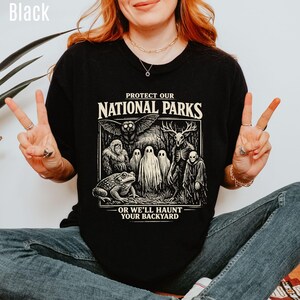 May include: A black t-shirt featuring a graphic print with the text PROTECT OUR NATIONAL PARKS OR WE'LL HAUNT YOUR BACKYARD. The design includes illustrations of a toad, a sasquatch, an owl, ghosts, a deer-skulled creature, and a hooded figure.