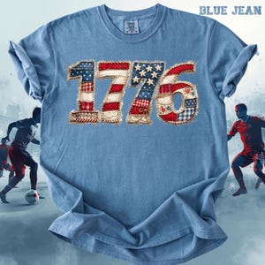May include: A light blue t-shirt with the number "1776" in a patchwork design featuring red, white, and blue patterns. The text "BLUE JEAN" is at the top right. The shirt has short sleeves and a crew neck.