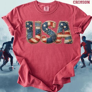 May include: A red t-shirt with the word "USA" appliquéd on the front. The letters are made of denim fabric with a patchwork design featuring the American flag. The shirt has a crew neck and short sleeves. The word "CRIMSON" is printed at the top.