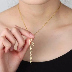May include: A gold necklace featuring a pendant spelling "Louise" in cursive script. The necklace is worn. The pendant is held by a hand.