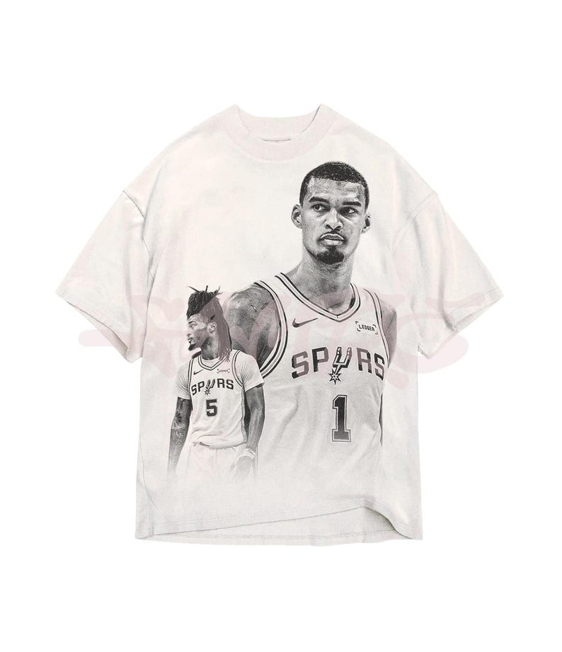 May include: White t-shirt with a black and white graphic of a basketball player in a Spurs jersey. The jersey displays the number 1 and the word "SPURS". The shirt has a crew neck and short sleeves.