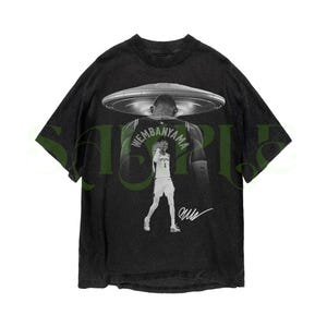 May include: Black graphic t-shirt featuring a basketball player with a UFO design. The player's name, "Wembanyama," is visible on the jersey. The shirt has a vintage, distressed look with a signature at the bottom.