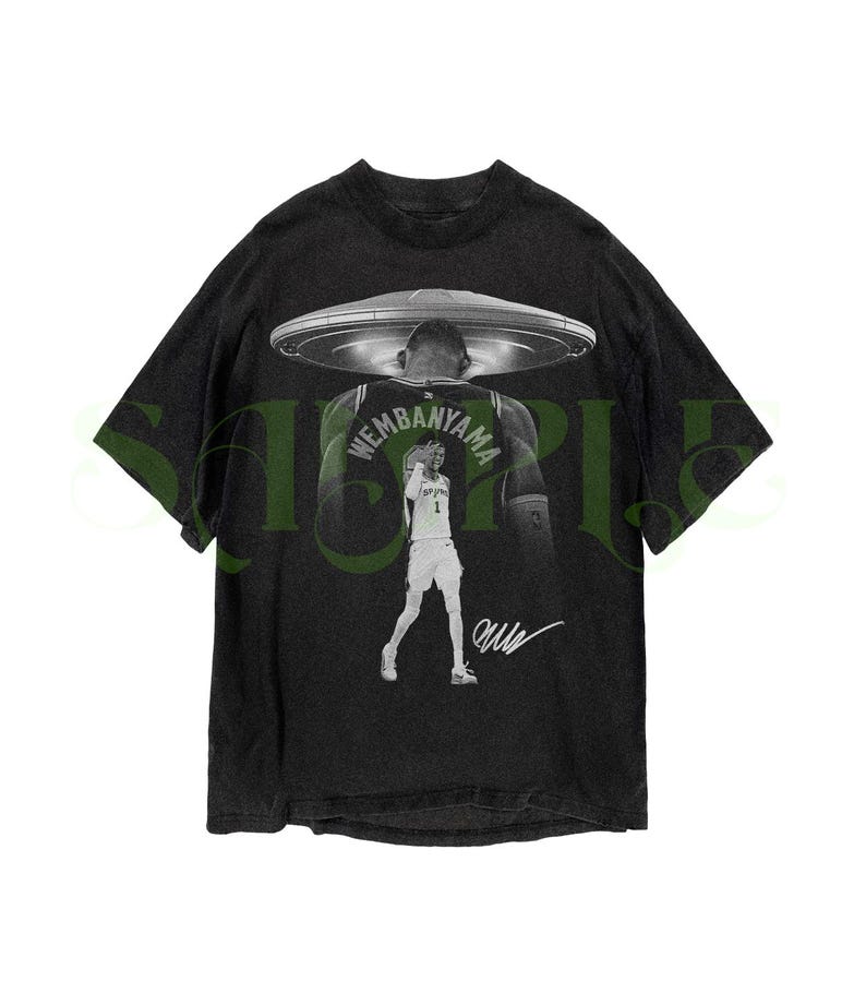 May include: Black graphic t-shirt featuring a basketball player with a UFO design. The player's name, "Wembanyama," is visible on the jersey. The shirt has a vintage, distressed look with a signature at the bottom.