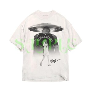 May include: White t-shirt featuring a black and white graphic of a basketball player with a UFO above his head. The player's jersey has the name "WEMBANYAMA" on the back. The shirt also includes a signature. The word "SAMPLE" is printed in green.
