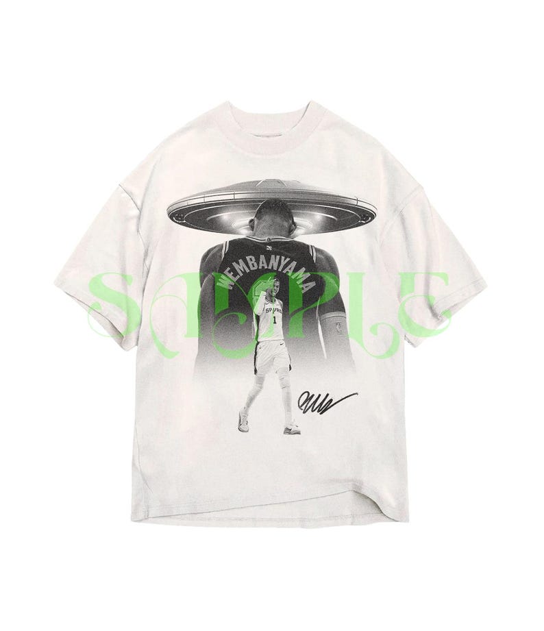 May include: White t-shirt featuring a black and white graphic of a basketball player with a UFO above his head. The player's jersey has the name "WEMBANYAMA" on the back. The shirt also includes a signature. The word "SAMPLE" is printed in green.