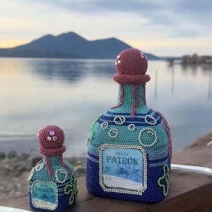 Może przedstawiać: Dwie dekoracyjne butelki tequili Patron, jedna duża i jedna mała, zdobione są misternymi koralikami w odcieniach turkusu, błękitu i różu. Butelki mają etykietę z napisem "Silver Patron".