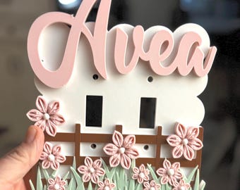 May include: A white light switch cover plate featuring the name Avea in pink script lettering at the top. The design includes a brown fence, light green grass, and several small pink fabric flowers with white centers arranged along the bottom.