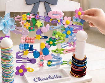 May include: A white wooden hair accessory organizer with a gray bow on top. The organizer holds various colorful hair clips, barrettes, and hair ties. A tray at the bottom is labeled "Charlotte".