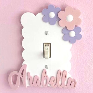 Op de afbeelding: Een witte lichtschakelaarafdekplaat met geschulpte randen en de naam Arabella in roze cursief schrift onderaan. De rechterbovenhoek is versierd met drie gelaagde bloemuitsneden in tinten lichtpaars en zachtroze.