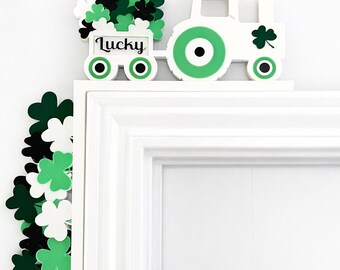 May include: A white tractor-shaped decoration with green and black accents, carrying a cart filled with shamrocks. The word "Lucky" is written on the cart. A shamrock border is on the left side. Perfect for St. Patrick's Day.