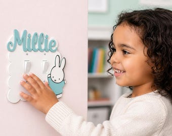 May include: A white light switch cover with a scalloped edge, featuring the name "Millie" in teal script and a cartoon bunny. The cover has two light switches and is mounted on a pink wall.