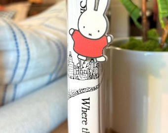 May include: A clear acrylic bookmark featuring a white cartoon bunny wearing a red shirt. The bookmark is attached to a book with the title "Where the Sea". The book has a black and white illustration of a city skyline.