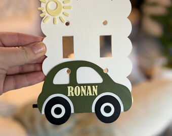 May include: A decorative light switch cover featuring a green car with black wheels and the name "RONAN" in gold lettering. The cover has a white background with a yellow sun design.