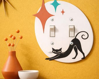 May include: An oval-shaped white double light switch plate featuring a black cat stretching its body across the bottom. The design includes a large orange four-pointed star and a smaller teal four-pointed star above the two toggle switches.