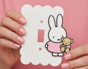 May include: White light switch cover with a scalloped edge, featuring a cartoon bunny in a pink dress holding a teddy bear. The bunny has black outlines and a black nose and eyes. The switch plate has a rectangular opening for the switch.