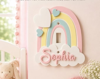 May include: A wall-mounted light switch plate shaped like a rainbow with pastel pink, yellow, and blue arches. The plate features a white cloud base with the name Sophia in pink script lettering and two white and blue heart accents attached.