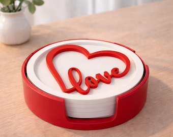 May include: A set of white coasters with a red heart and the word "Love" in a cursive font. The coasters are in a red holder. The set is on a light brown surface.