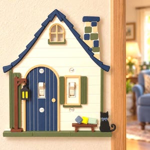 May include: A house-shaped light switch cover featuring a cream-colored facade, a dark blue roof, and a blue arched door. It includes a chimney, a hanging lantern, a small bench with three cushions, and a black cat figure on the side.