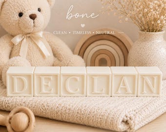 May include: Six cream-colored square blocks arranged in a row, each featuring a raised letter spelling the name DECLAN. The blocks are positioned on a textured cream blanket in front of a plush teddy bear, a wooden rainbow, and a speckled vase.