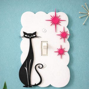 May include: A light switch cover with a black cat design and pink starburst decorations. The white cover has a scalloped edge, and the background is light blue. The switch is a standard size, suitable for UK homes.