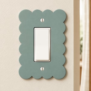 May include: A sage green light switch wall plate featuring a scalloped edge design. The rectangular plate has a central vertical opening for a rocker switch, with two visible silver screws securing it to the wall surface.