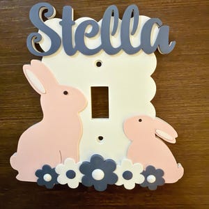 Op de afbeelding: Een witte lichtschakelaarplaat met de naam Stella in blauwe schrijfletters aan de bovenkant. De plaat is versierd met twee lichtroze konijnen en een rij afwisselend blauwe en witte bloemen langs de onderrand van de rechthoekige afdekplaat.