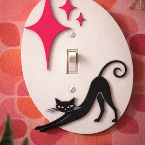 Puede incluir: Una cubierta decorativa para interruptor de luz con un gato negro estirándose sobre un óvalo blanco. La cubierta incluye un interruptor de luz y está adornada con acentos de estrella rosa. El fondo es un patrón floral rosa y rojo.