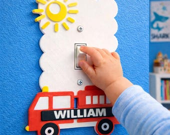 May include: A decorative light switch cover featuring a red fire truck with the name "WILLIAM" in white letters. The cover includes a yellow sun and cloud design. A child's hand is reaching for the light switch on a blue wall.