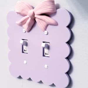 Puede incluir: Una placa de pared para interruptor doble de color violeta claro con borde festoneado y un lazo rosa claro tridimensional fijado en la parte superior. La placa cuenta con dos interruptores blancos y se fija a la pared con cuatro tornillos plateados.