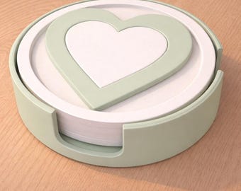 May include: A set of coasters with a heart design. The coasters are white with a light green heart in the center and a light green border. The coasters are stacked in a matching light green holder. The set is on a wooden surface.