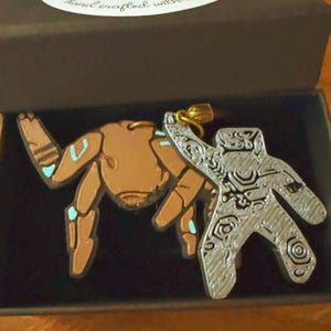 Peut inclure: Deux pendentifs dans une boîte noire. L'un est en forme de crabe couleur cuivre avec des accents bleu clair. L'autre est une figurine couleur argent avec un motif texturé en tourbillons. Les deux pendentifs ont des anneaux en métal doré.