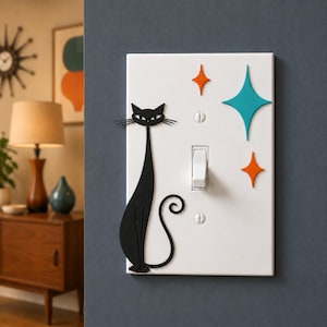 May include: A white rectangular light switch cover plate featuring a black cat silhouette on the left side and three colorful starburst shapes in orange and teal on the right side, secured with two white screws above and below the switch.