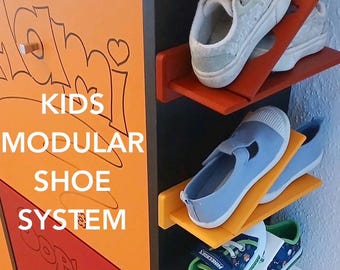 May include: A modular shoe storage system featuring a vertical black frame with orange, yellow, and white angled shelves. The unit displays the text KIDS MODULAR SHOE SYSTEM on an orange panel. Three pairs of children's shoes are placed on the shelves.