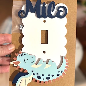Op de afbeelding: Een witte, geschulpte lichtschakelaarafdekplaat met de naam Milo in donkerblauw schrift bovenaan en een lichtblauwe draak met donkerblauwe stippen en oranje stekels onderaan. De plaat bevat twee schroefgaten en een zakje met schroeven.