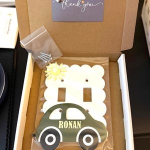 Op de afbeelding: Een witte dubbele lichtschakelaarplaat met een geschulpte rand, een gele zon en een donkergroene auto-afbeelding met de naam RONAN in het geel. Het item zit in een kartonnen doos met een bedanksticker en een zakje met montageschroeven.