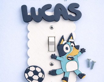 May include: A white scalloped light switch plate featuring the name LUCAS in dark blue letters at the top, a blue cartoon dog character, and a black and white soccer ball. The plate includes two metal screws and text reading Includes Matching Screws.