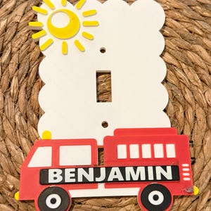 Op de afbeelding: Een decoratieve lichtschakelaar cover met een rode brandweerwagen met de naam "BENJAMIN", een gele zon en een witte wolk-vormige achtergrond. De schakelaar cover heeft een standaard tuimelschakelaar opening.