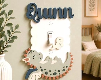 May include: A white scalloped light switch cover featuring the name Quinn in dark blue script at the top. A light green cartoon dragon with dark green spots and orange spikes lies across the bottom, with a small white cloud near the switch.