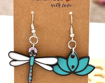 May include: A pair of handmade earrings featuring a dragonfly and a lotus flower design. The dragonfly has white wings, a turquoise body, and purple accents. The lotus flower is turquoise with black outlines. The earrings are on a brown card with the text "Handmade with love".