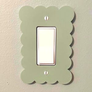 Op de afbeelding: Een saliegroene afdekplaat voor een lichtschakelaar met een geschulpt randontwerp. De rechthoekige plaat is bevestigd met twee zichtbare zilverkleurige schroeven en omringt een standaard witte rechthoekige wipschakelaar.