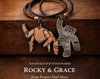 May include: Two bookmarks on a dark brown leather cord. One pendant is a tan, multi-legged creature with light blue accents. The other is a textured, silver-toned metal piece with intricate patterns. Text reads: HANDCRAFTED BOOKMARKS ROCKY & GRACE from Project Hail Mary.
