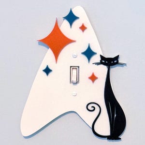 May include: A white, boomerang-shaped light switch cover plate featuring a black cat silhouette on the right side. The plate is decorated with orange and blue four-pointed star shapes of varying sizes scattered across the white surface.