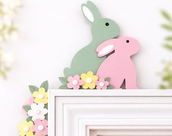 May include: A decorative Easter-themed wooden display featuring two pastel-colored bunnies, one green and one pink, with white ears. The bunnies are surrounded by yellow, pink, and white flowers with green leaves. The display is placed on a white surface.