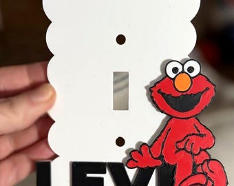 May include: A white, scalloped-edge light switch cover featuring a red Elmo character and the name "LEVI" in black letters. The Elmo figure has a black mouth, white eyes, and an orange nose.